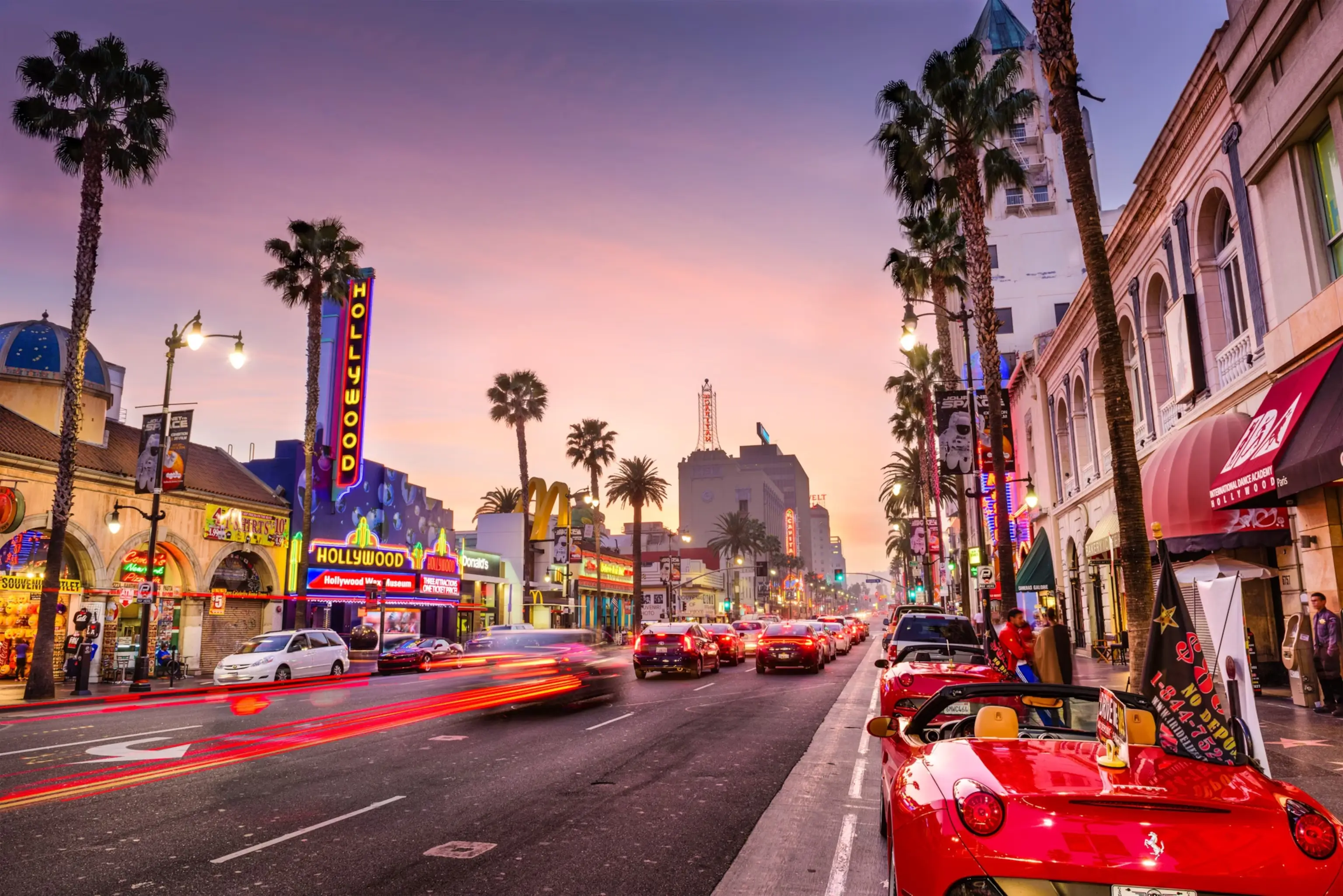 Vibrant sunset view of Hollywood Boulevard in Los Angeles featuring neon signs, palm trees, and a red convertible Ferrari parked on the street.