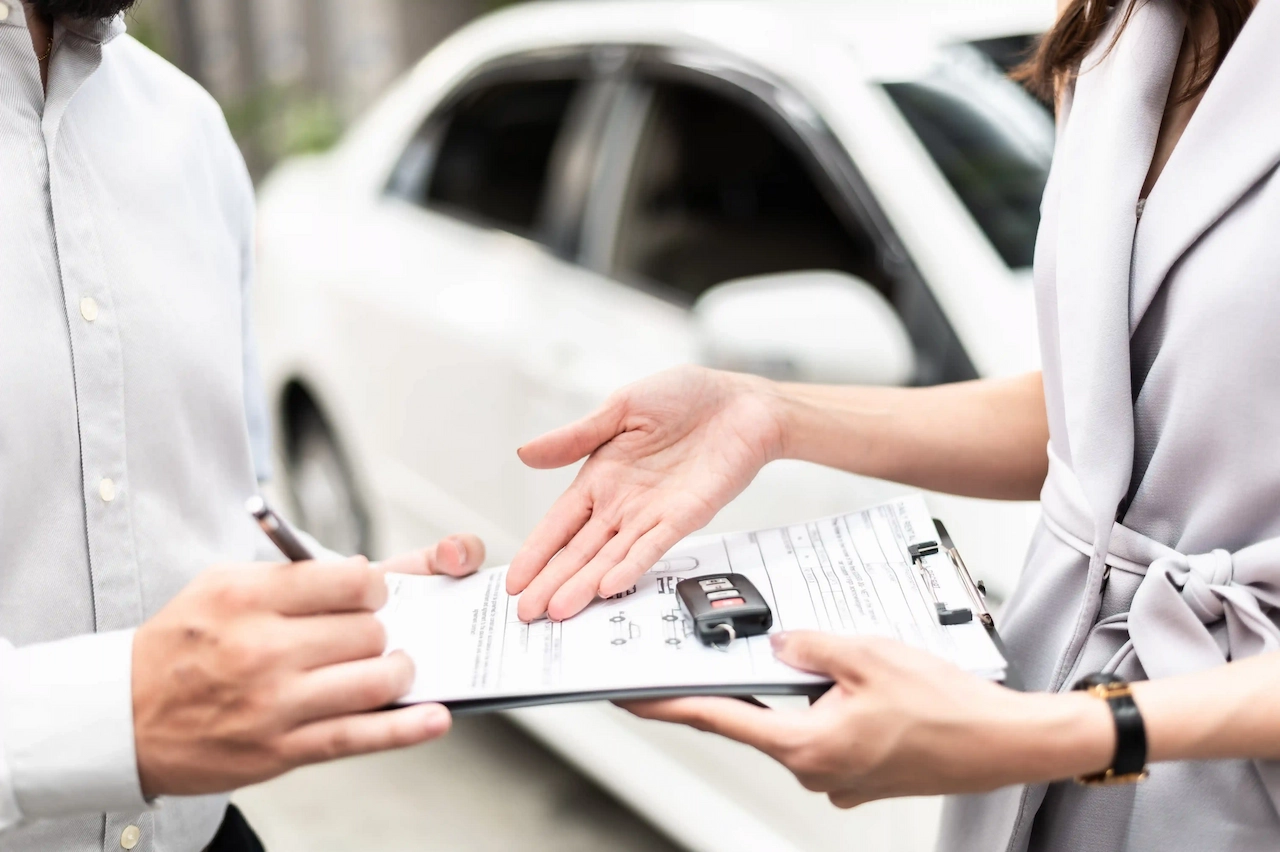 A close-up view of a customer using a pen to sign a car hire agreement on a clipboard, which also holds a set of car keys, handed to them by a representative with a white vehicle blurred in the background.
