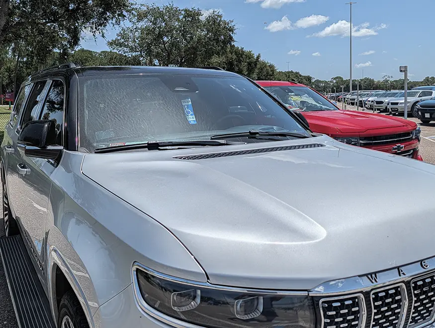 Florida accessible parking space showing vehicle and striped access aisle demarcation Silver Jeep parked in a Florida blue-marked accessible parking space with clearly visible white-striped access aisle beside the vehicle. Background shows grass, trees and a second red vehicle; daytime outdoor lot.
