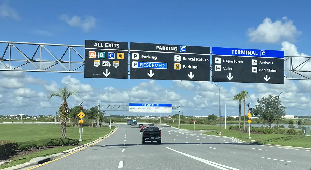 Overhead road gantry signage at Orlando International Airport Terminal C directing drivers toward rental car return lanes and parking facilities under a blue sky in Florida.