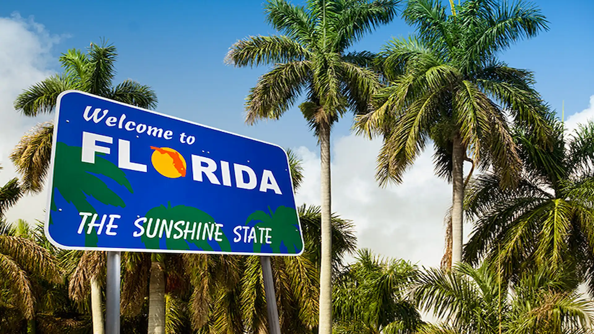 A blue highway road sign reading 'Welcome to Florida THE SUNSHINE STATE' surrounded by tall green palm trees against a partly cloudy sky, marking the beginning of a Florida road trip.