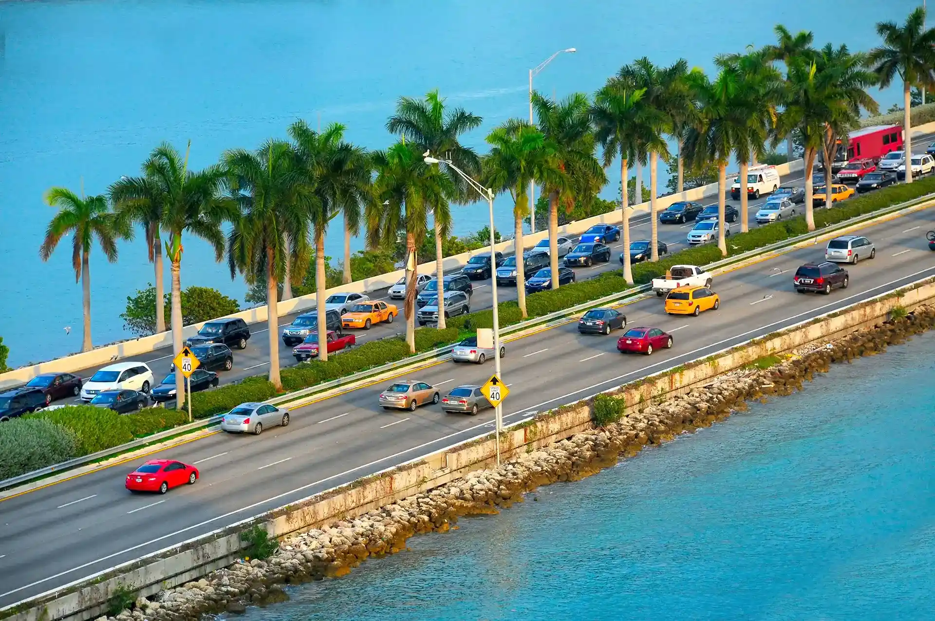 An elevated view of cars driving along a multi-lane coastal highway lined with tall palm trees and surrounded by bright blue water, illustrating a scenic Florida road trip.