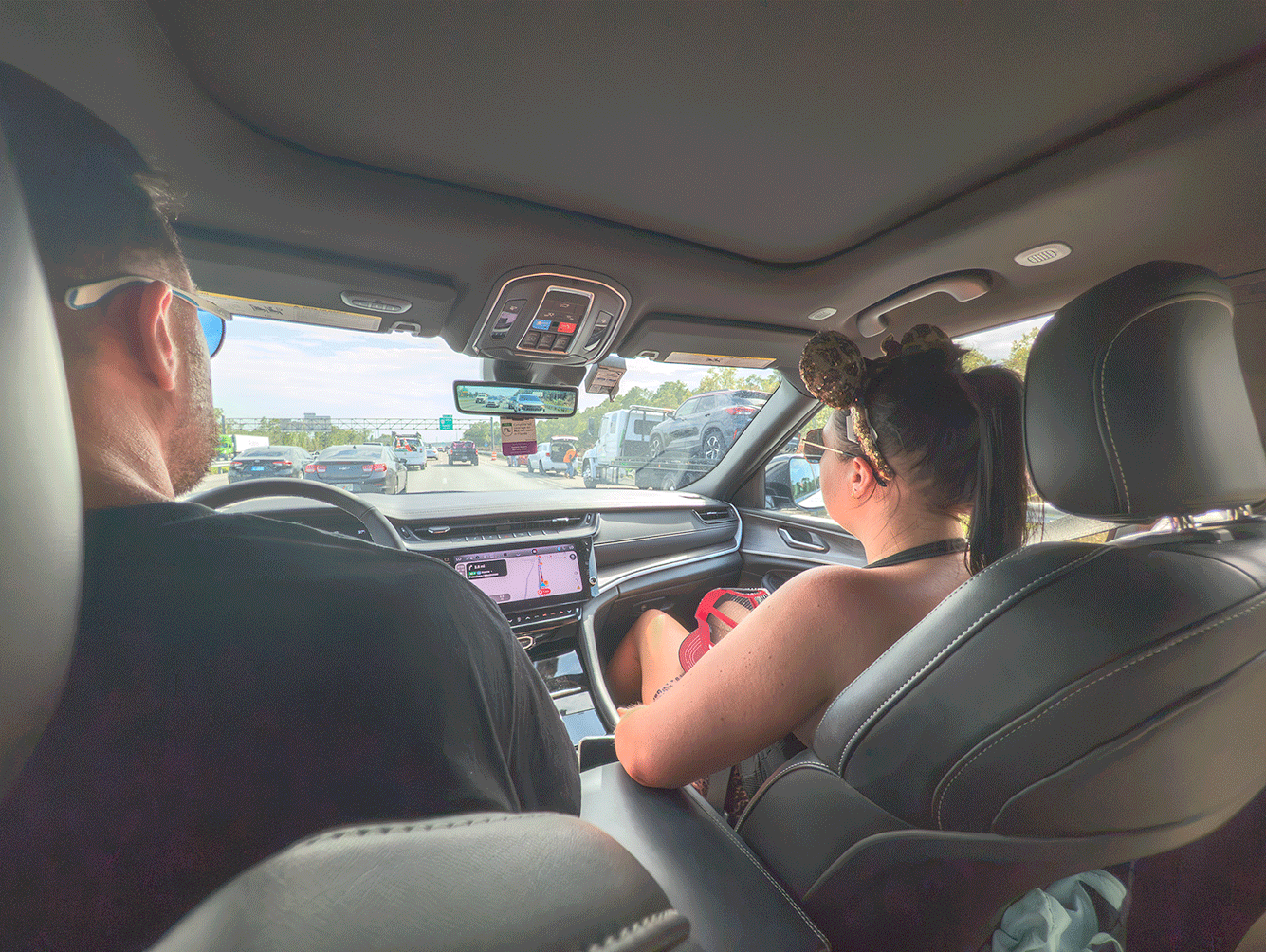 Couple driving in a modern rental car on a sunny highway, with the passenger wearing leopard-print Minnie Mouse ears.