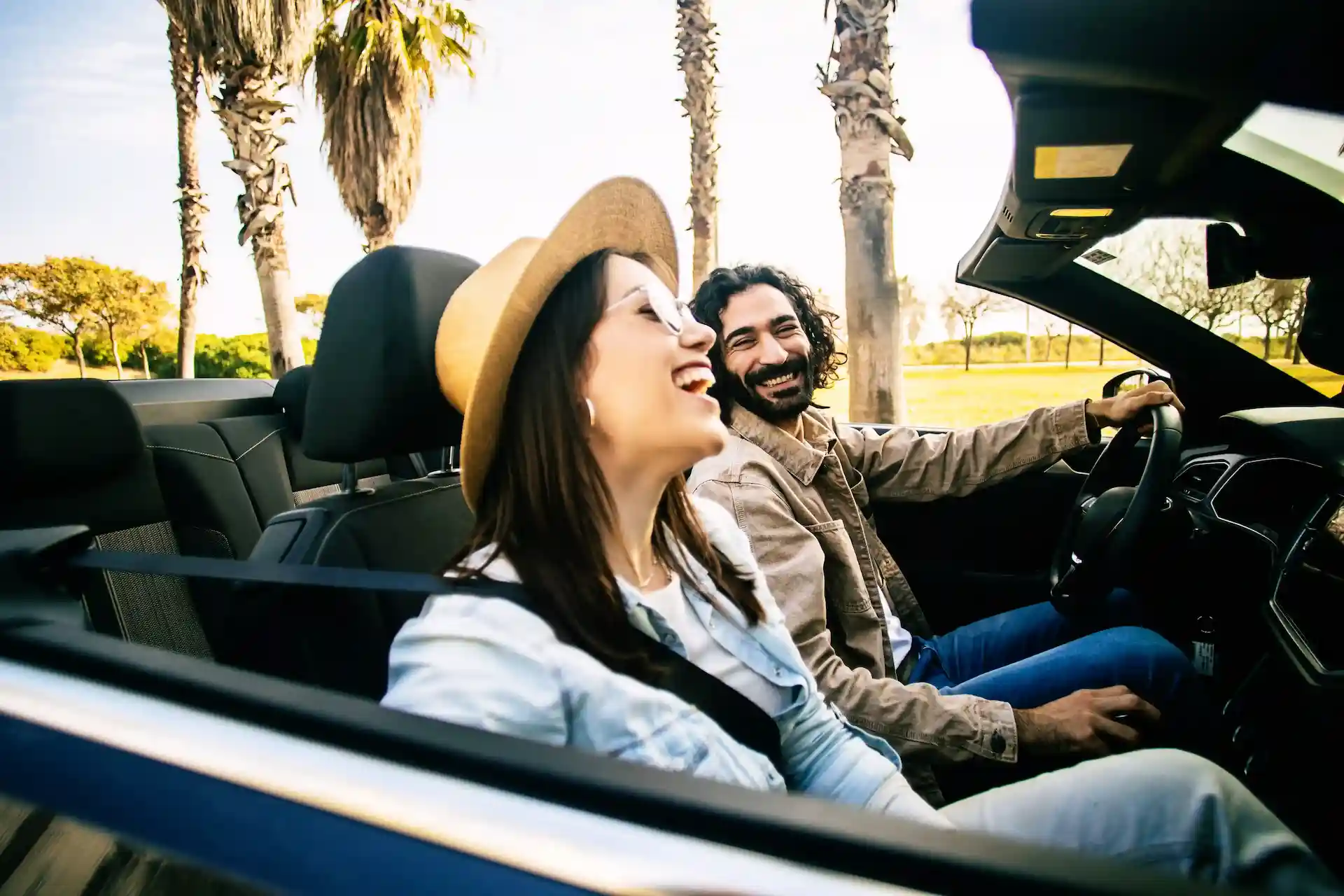 A laughing woman wearing a straw hat and a smiling man driving a dark convertible hire car with the roof down past Florida palm trees in the bright sunshine.