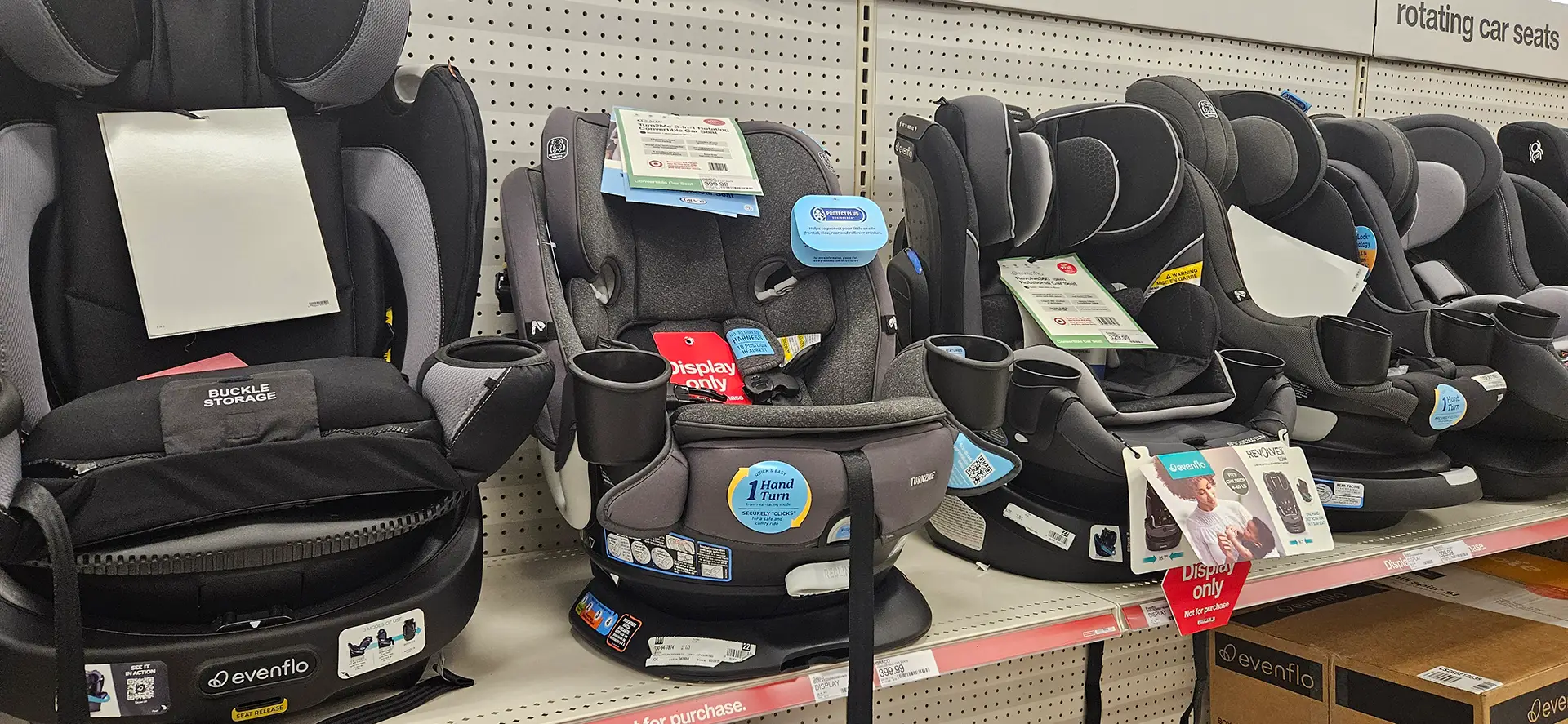 A row of grey and black child car seats, including brands like Evenflo, sitting on display shelves in an American retail store with 'Display only' tags hanging from the harnesses.