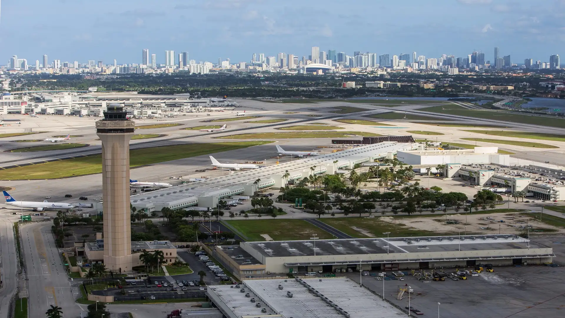 Aerial view of Miami International Airport (MIA) showing the main control tower, runways, and terminal buildings, with the downtown Miami skyline in the background, representing the arrival point for UK holidaymakers collecting their hire car.