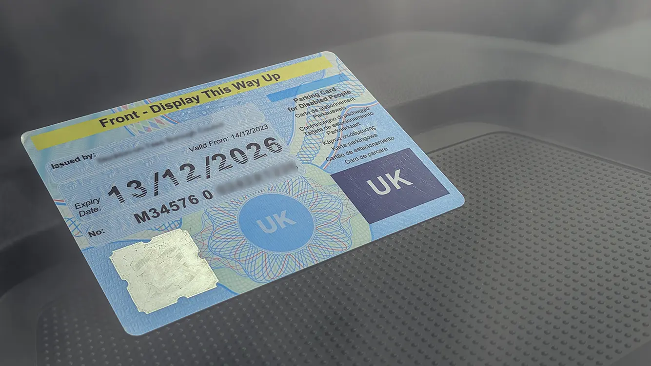 UK Blue Badge correctly displayed face-up on a rental car dashboard, clearly visible through the windscreen. Badge shows "Front - Display This Way Up" instruction, valid expiry date, and UK marking.